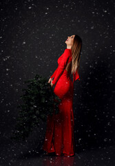 Young glamour woman walking in long red dress with winter christmas fir tree under heavy snow. New year concept background