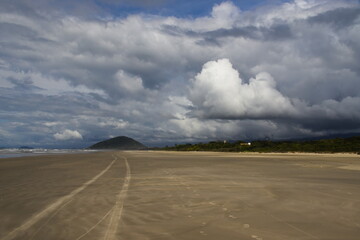 tire tracks and footprints in the golden sand of the beach, converging on a small hill among the vegetation, under a blue sky covered by large shaded cumulus clouds.