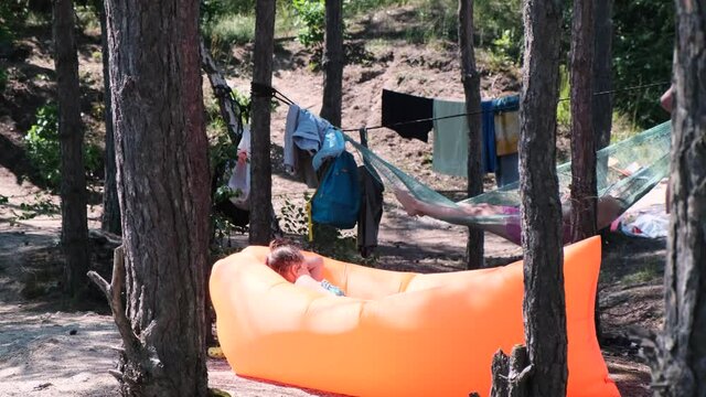 Mental health, camping, outdoor recreation. Young woman lies on an orange inflatable sofa or couch among the trees. Man lies on hammock in the background.