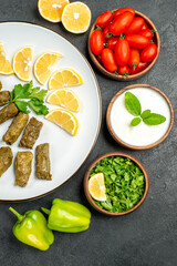 top view stuffed grape leaves parsley leaves and lemon half slices on plate bowls with cherry tomatoes natural yogurt green peppers slices of lemon on dark background