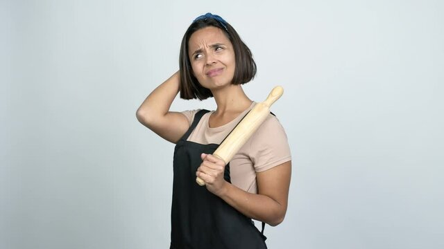 Young latin woman holding a rolling pin having doubts while scratching head over isolated background