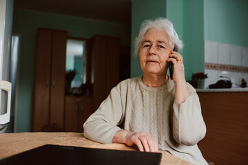 Portrait of a senior grandmother talking on the phone with her family while sitting at a table in the house. Communication during isolation of COVID-19 coronavirus