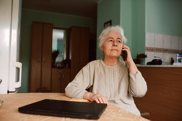 A senior grandmother talking on the phone with her family while sitting at a table in the house. Communication during isolation of COVID-19 coronavirus