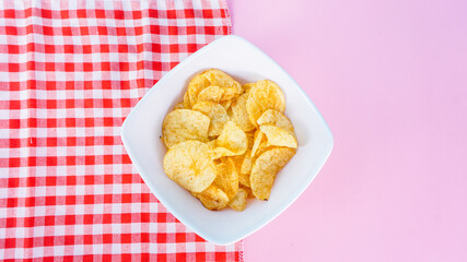 Potato chips in bowl on napkin and pink background, top view. Salty crisps scattered on a table