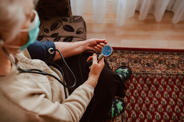 Close-up of a senior woman with a face mask that measures pressure with a pressure gauge while sitting on the couch. Health check during the COVID-19 coronavirus pandemic