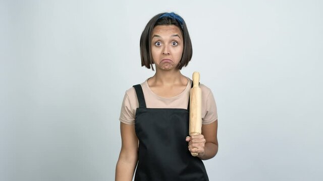 Young latin woman holding a rolling pin making unimportant and doubts gesture while lifting the shoulders and the palms of the hands over isolated background