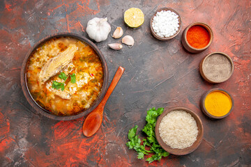 top view homemade chicken and rice soup in a bowl wooden spoon bowl of rice garlic slice of lemon parsley leaves different spices on dark red background