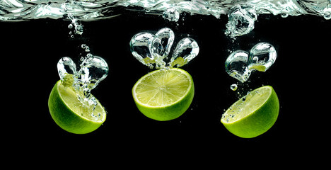 Fototapeta na wymiar Bunch of lime fruits halves splashing with bubbles into water isolated against black background. Citrus theme, panorama