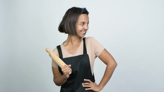 Young latin woman holding a rolling pin posing with arms at hip and laughing over isolated background