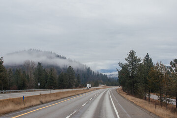 Fototapeta premium Highway with road signs on the sides among high mountains in the clouds in winter, along which trucks and cars travel. Idaho, USA, 12-5-2020