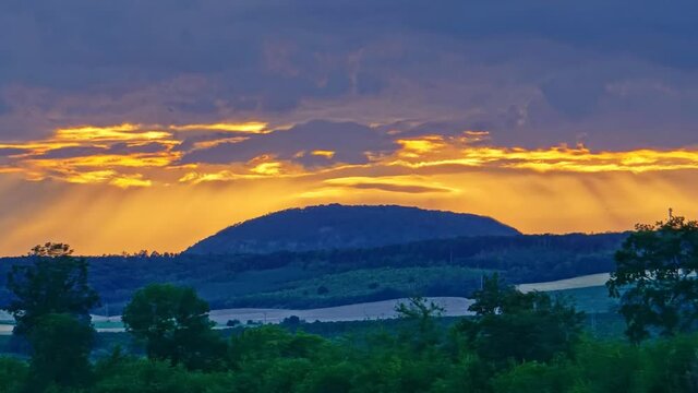 Hill under moving clouds illuminated by sun at sunset. Trees and green plants are in the dark foreground, sun rays shine through the clouds and illuminate them to yellow around the hill. Czechia