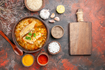 top view homemade chicken and rice soup in a bowl wooden spoon bowl of rice garlic slice of lemon different spices a cutting board on dark red background
