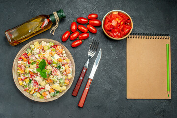 top view of vegetable salad with tomatoes oil bottle cutleries and notepad on side on dark grey background