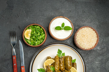 top view stuffed grape leaves on white plate bowls of rice natural yogurt parsley knife and fork on dark background
