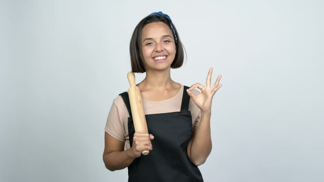 Young latin woman holding a rolling pin showing ok sign with fingers over isolated background