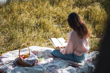 Back view of a long-haired woman reading a book sitting in the grass having a picnic in nature...