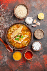 top view homemade chicken and rice soup in a bowl wooden spoon bowl of rice garlic slice of lemon different spices on dark red background