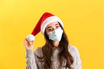 Fototapeta premium Beautiful woman wearing Santa Claus hat and medical mask on yellow background