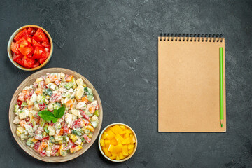 top view of bowl of vegetable salad on left bottom side with bowls of vegetables and notepad on side on dark green-grey background