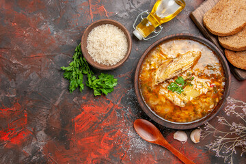 top view chicken and rice soup in a bowl wooden spoon garlic bottle of oil parsley bowl of rice slices of bread on chopping board on dark red background with copy space