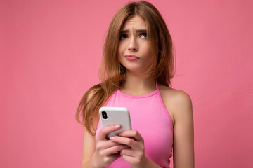 Closeup photo of beautiful thoughtful young blonde woman wearing pink top poising isolated on pink background with empty space holding in hand and using mobile phone surfing on the internet online