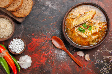 top view homemade chicken and rice soup in a bowl wooden spoon hot peppers bowls with sea salt and rice garlic slices of bread on chopping board on dark red background