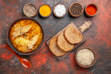 top view bowl of chicken soup bowls with different spices wooden spoon slices of bread on cutting board bowl of rice on dark red background