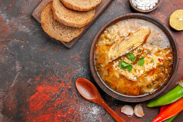 top close view chicken and rice soup in a bowl wooden spoon hot peppers bowl with sea salt garlic slice of lemon slicesof bread on cutting board on dark red background with copy space