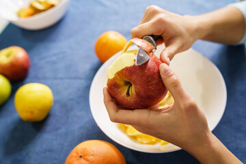 Close up on hands of unknown caucasian woman peeling red apple over the bowl on the table at home -...
