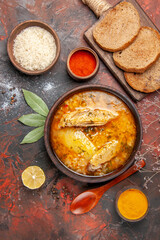 top view chicken soup bowl with rice slice of lemon bread slices on cutting board red pepper powder turmeric wood spoon on dark red background