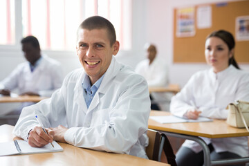 Portrait of cheerful man in white coat sitting in lecture hall on refresher course, looking at camera with smile