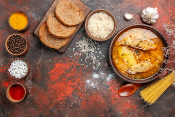 top view bowl of chicken soup garlic bowl of rice dry spaghetti slices of bread on cutting board spoon bowls with different spices on dark red background with copy space