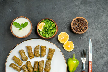 top view stuffed grape leaves on plate bowls with natural yogurt black pepper parsley green pepper slices of lemon fork and knife on dark background with copy space