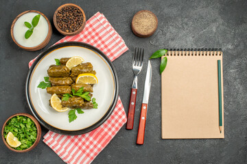 top view stuffed grape leaves on white plate bowls of natural yogurt and black pepper knife and fork napkin salt a notebook on dark background