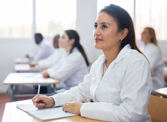 Positive latin american girl in white coat listening to lecture and taking notes in classroom...