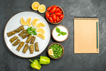 top view stuffed grape leaves parsley leaves and lemon half slices on plate bowls with cherry tomatoes natural yogurt green peppers slices of lemon a notebook on dark background