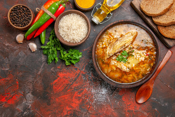 top close view chicken and rice soup in a bowl wooden spoon hot pepper bottle of oil parsley bowl of rice slices of bread on chopping board different spices on dark red background with copy space