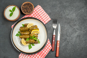 top view stuffed grape leaves on white plate bowls of natural yogurt and black pepper knife and fork napkin on dark background with copy space