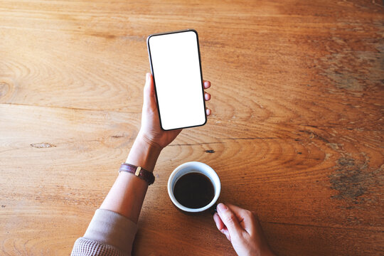 Top view mockup image of a woman holding mobile phone with blank white desktop screen while drinking coffee