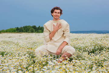 Fototapeta na wymiar Tall handsome man sitting on a back of a chair in camomile flowers field