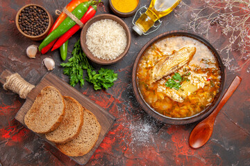top view chicken and rice soup in a bowl wooden spoon hot pepper bottle of oil parsley bowl of rice slices of bread on chopping board different spices on dark red background