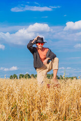 Naklejka na ściany i meble Tall handsome man with black hat and pink sunglasses standing with one leg on a brown vintage leather suitcase at golden oat field.