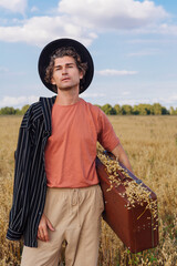 Tall handsome man holding brown suitcase full of ears of oats at golden oat field.