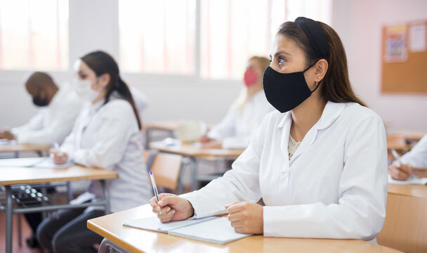 Interested Female Medical Student Wearing Protective Face Mask Making Notes During Group Lecture In Classroom. Necessary Precautions In Public Place During Coronavirus Pandemic