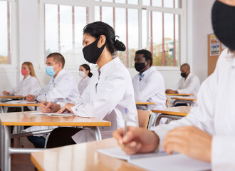 Fototapeta premium Adult asian female medical student in protective face mask studying in classroom with group, writing lectures in workbook. Concept of preventing spread of viral infections in pandemic