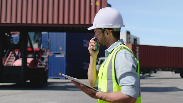 Foreman or cargo container worker walk along and in front of crane with container tank and also use walkie talkie to communicate with co-worker in workplace area.