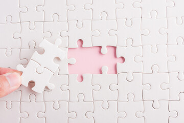 Top view flat lay of hand-holding last piece white paper jigsaw puzzle game last pieces put to place for solve problem complete mission, studio shot on a pink background, quiz calculation concept