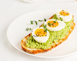 Avocado toast with toasted bread soft-boiled eggs with yellow yolk and tomatoes with herbs on white plate on light tablecloth