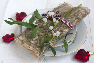 table setting: plates, a napkin tied with a ribbon, mistletoe branches, red glass Christmas toys, gingerbread cookies on a white wooden background. New Year, Christmas.