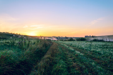 Tranquil summer rural landscape with road passing through farmlands during sunrise.Lush wet grass growing on meadow.